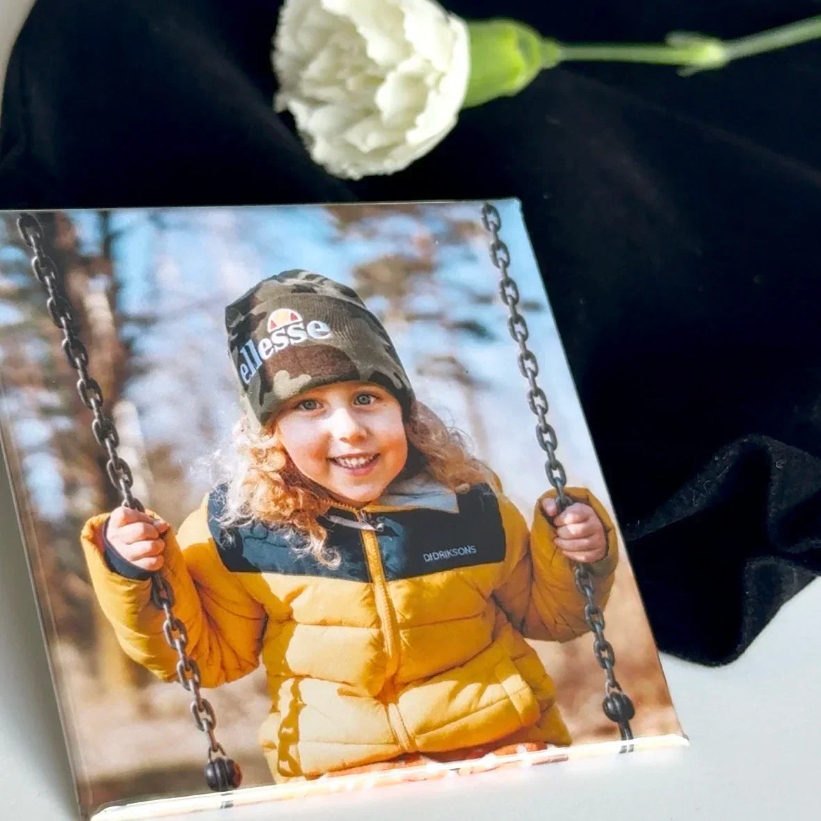 Square custom photo magnet displayed with gift bag and single white flower, displaying a nostalgic childhood memory of a girl on a swing.