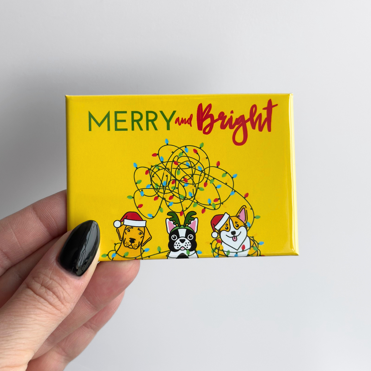 Hand holding a yellow novelty button magnet with Christmas-themed design and 'Merry and Bright' text. The magnet features an illustration of 3 dogs with tangled Christmas lights.