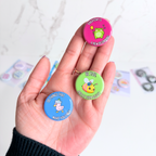 Hand holding three colorful button magnets with motivational phrases and character illustrations for size reference. Vivid prints and a glossy finish. 