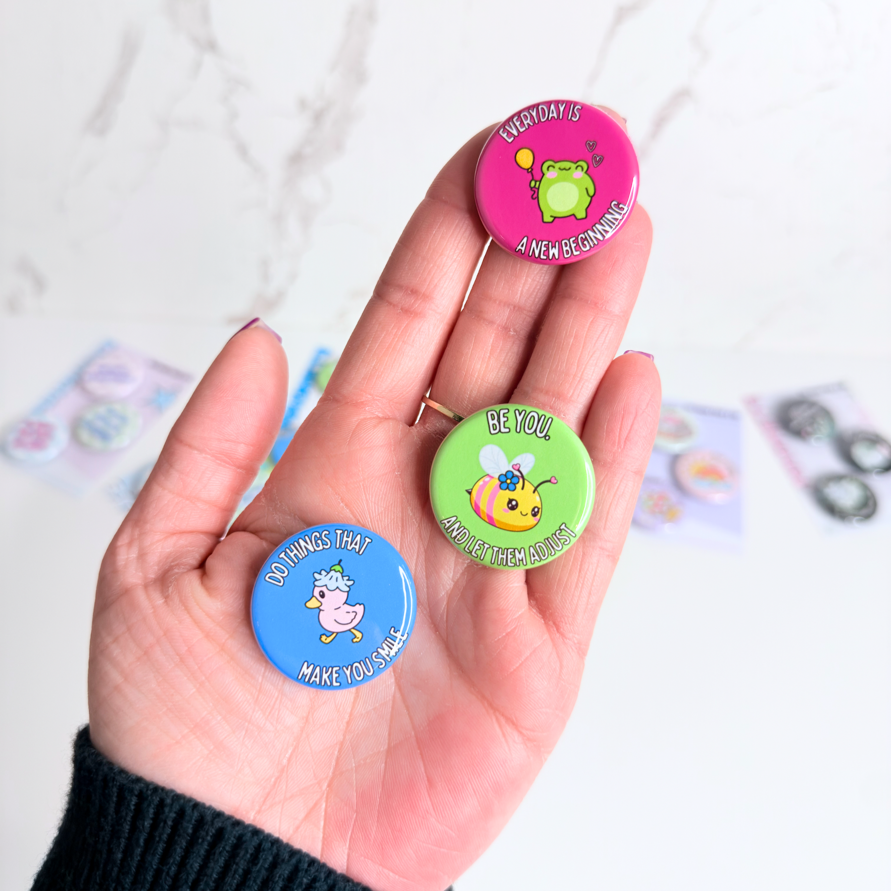 Hand holding three colorful button magnets with motivational phrases and character illustrations for size reference. Vivid prints and a glossy finish. 