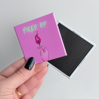 Hand holding bold pink novelty fridge button magnet with neon inspired text that reads 'fired up' and an illustration of a hand holding a lit match.