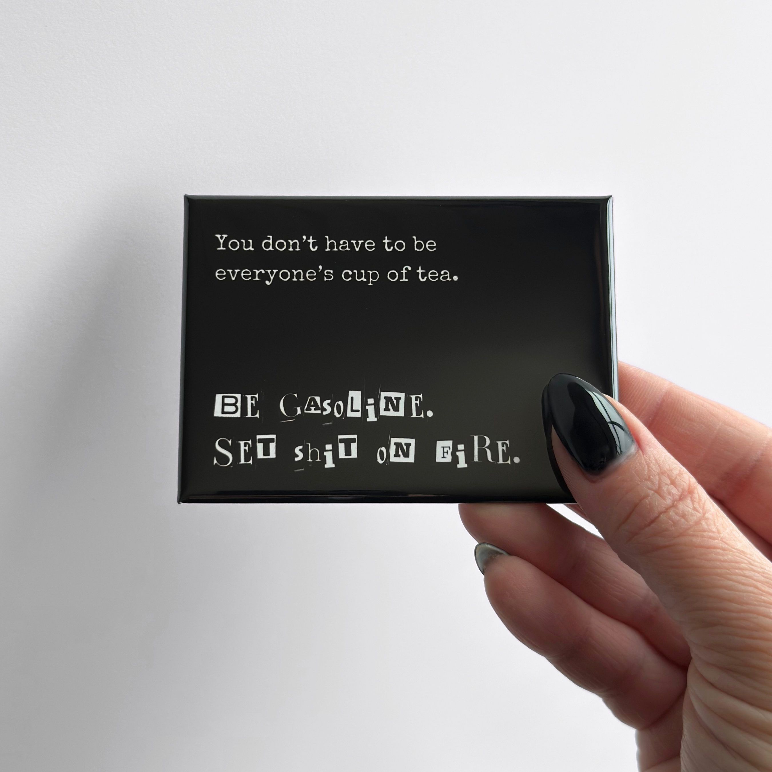 Hand holding a sarcastic novelty fridge button magnet shown in a rectangular format with a neutral gray background.