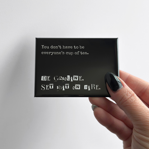 Hand holding a sarcastic novelty fridge button magnet shown in a rectangular format with a neutral gray background.
