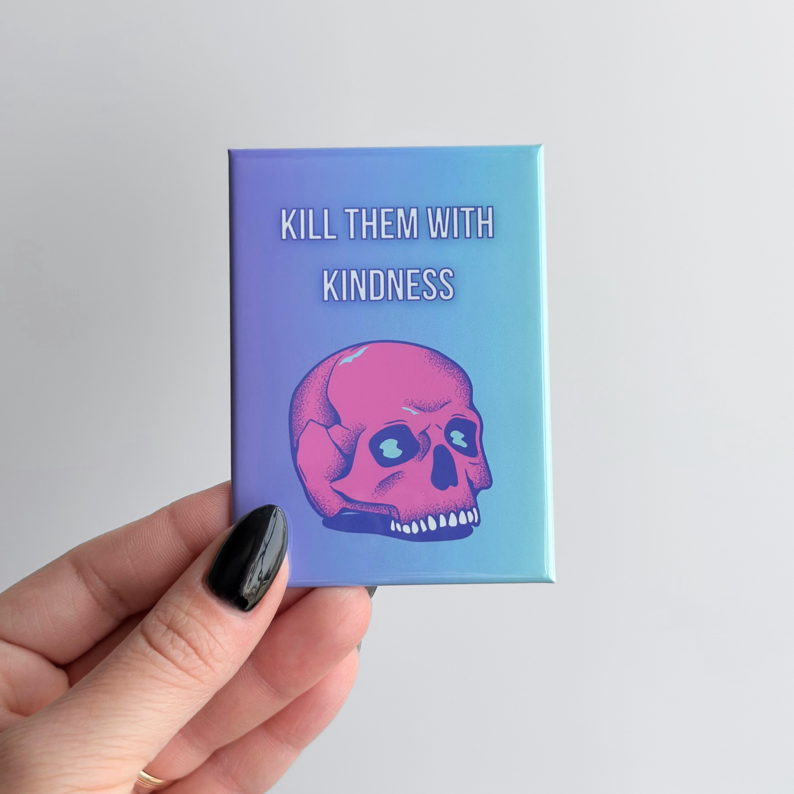 Dark humor novelty fridge magnet with a pink skull design and text 'Kill Them With Kindness' held by a hand against a light gray background.