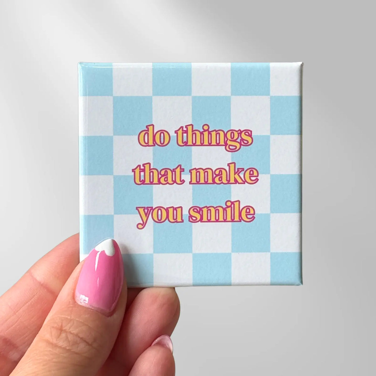A hand holds a small square magnet featuring a light blue and white checkerboard pattern with the text 'do things that make you smile' quote.