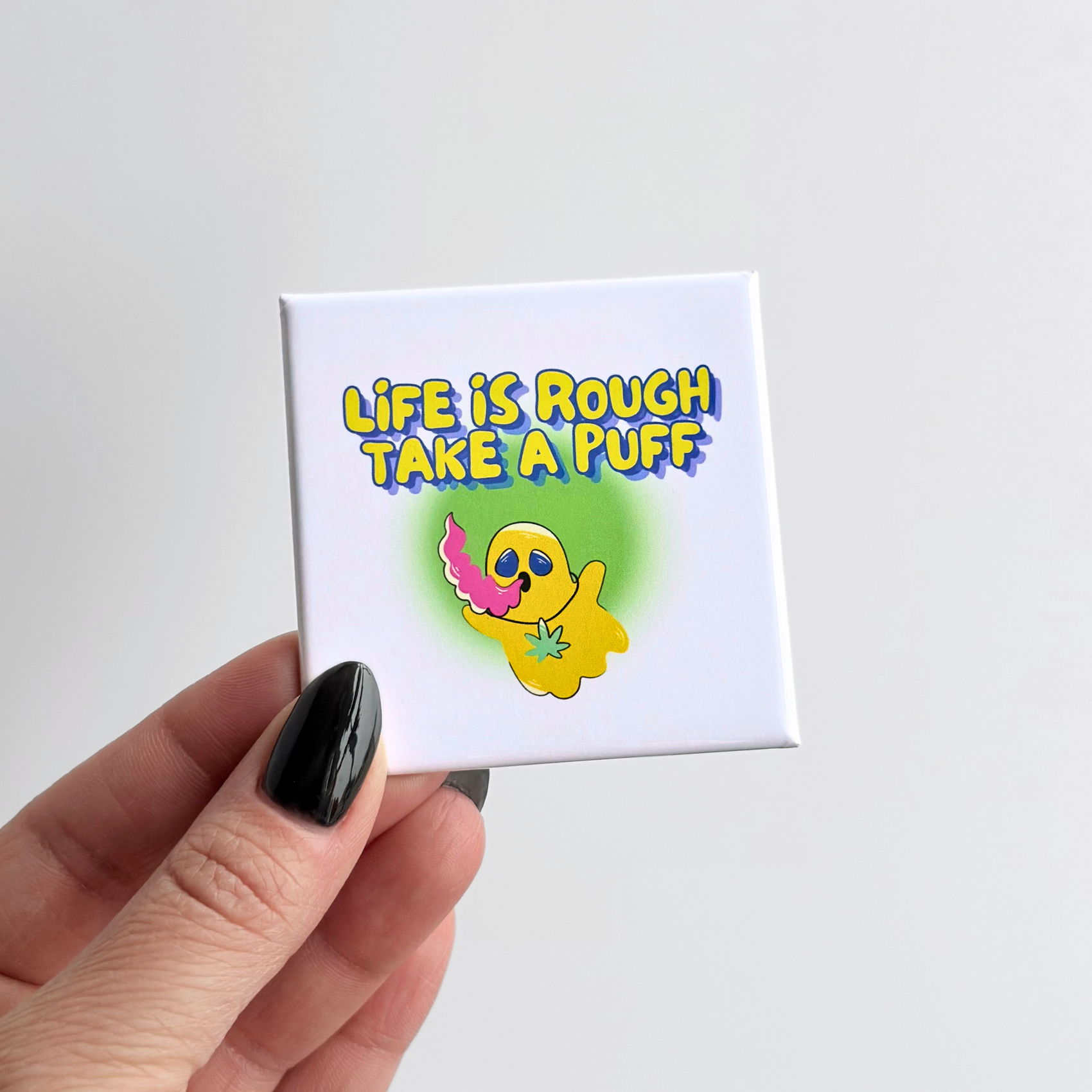 Hand holding a funny cannabis culture novelty fridge button magnet featuring a 'life is rough, take a puff' design with a floating ghost. 