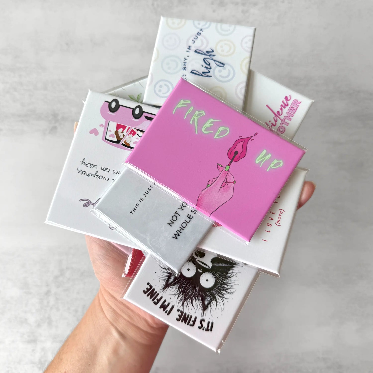 Hand holding a pile of ink and magnets novelty fridge magnets from each collection. Each magnet is unique and designed in house for unique gift giving or wholesale.