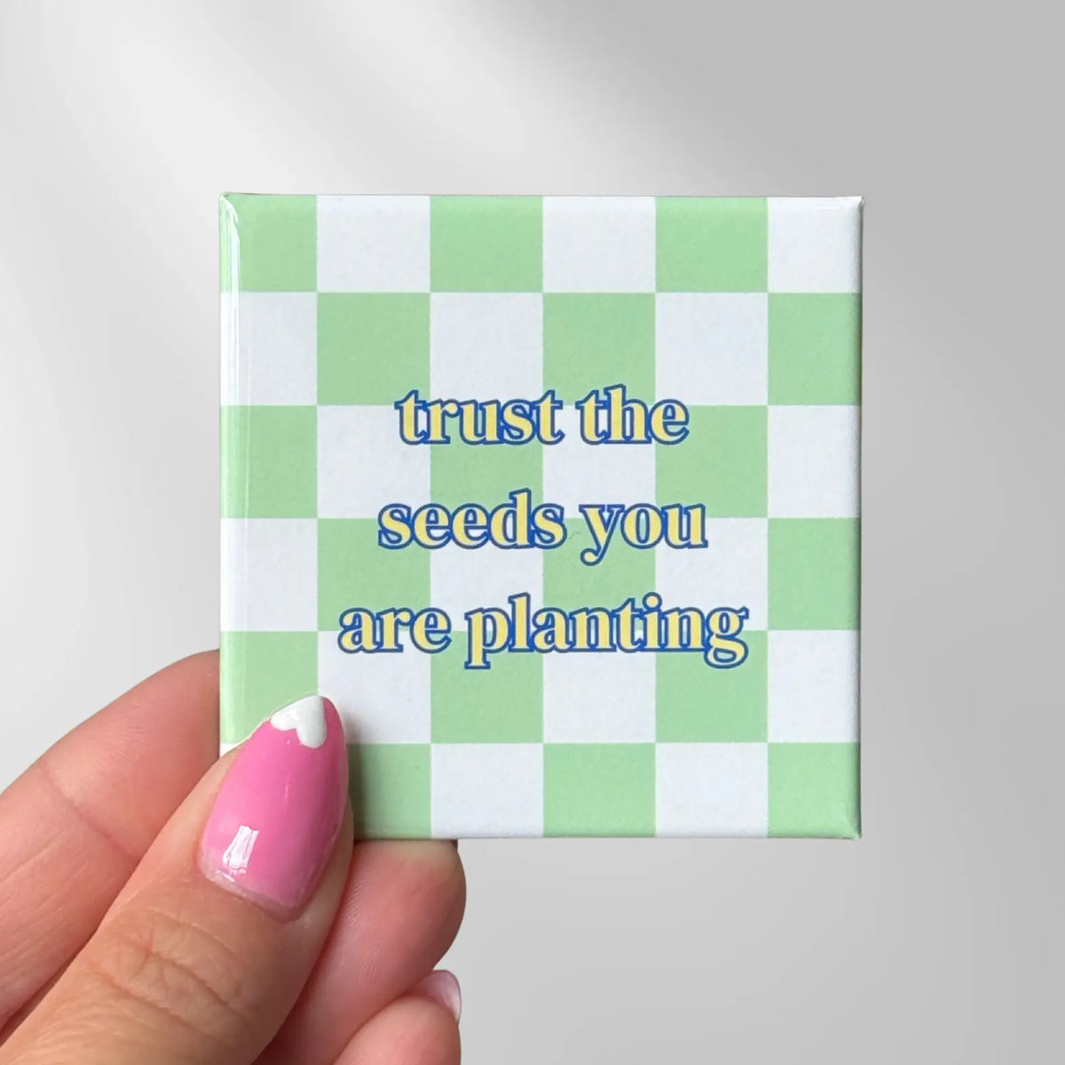 A hand holding a square magnet featuring a light green and white checkerboard pattern with the text 'trust the seeds you are planting' in blue outlined lettering. Maximalist decorative locker or fridge magnet.